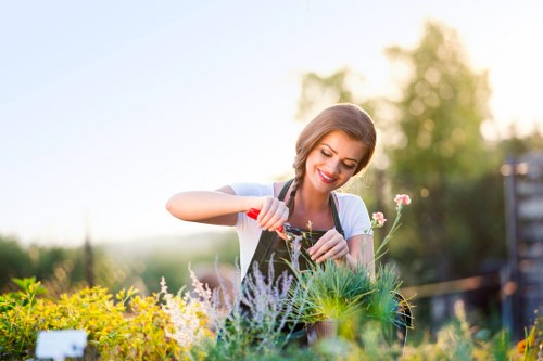 A young woman with a cheerful expression is tending to a variety of flowering plants and greenery in a well-maintained outdoor garden, with a background of blurred trees and a soft, natural light indicating a sunny day. In the foreground, there are lush green grass and flower beds containing different plants, some of which are flowering with small blooms in shades of pink, white, and yellow. The garden features a mix of shrubbery and flowering borders, with a wooden fence visible on the right side. The overall landscape suggests a cared-for outdoor space typical of residential gardens in the Eltham area, used for leisure and gardening activities. The scene highlights the natural, vibrant colours of the plants and the neat arrangement of garden elements, consistent with professional gardening and landscaping services offered by Gardening Services Eltham in the local area.