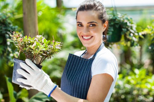 A young woman with dark hair tied back, smiling, wearing a navy and white striped apron over a white shirt and white gardening gloves, holding a potted shrub with small green leaves and light pink flowers in a lush garden setting with various green plants, trees, and a wooden post in the background. The scene appears to be outdoors on a bright, sunny day, with natural light illuminating the scene, reflecting a well-maintained garden environment suitable for gardening and landscaping services in Eltham.