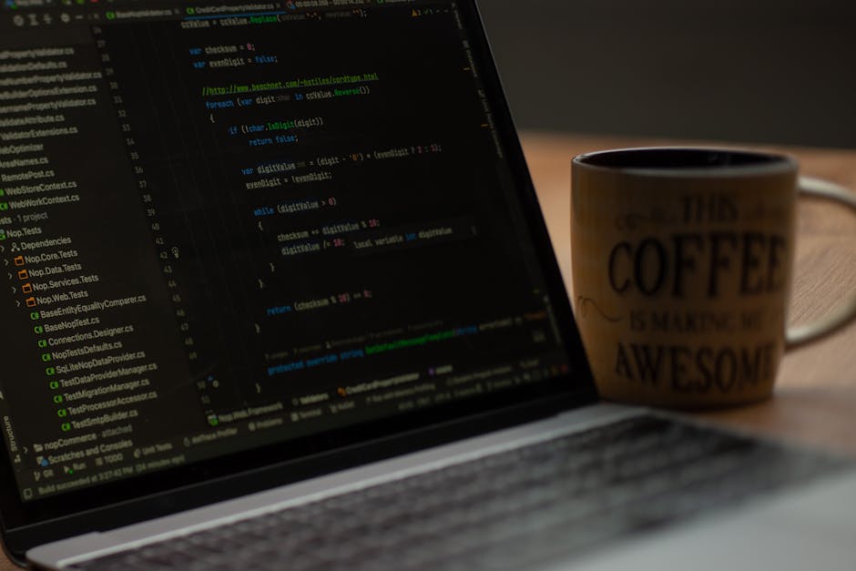 A close-up view of a laptop displaying lines of code on its screen, with a black coffee mug placed next to it on a wooden desk. The coffee mug has an off-white background with black text that reads 'T