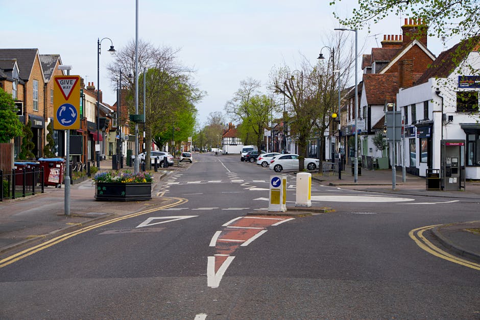 The image depicts a quiet urban street scene with a central traffic island featuring a red brick section and white dashed lines, separating lanes for vehicles. On either side, there are small shops an