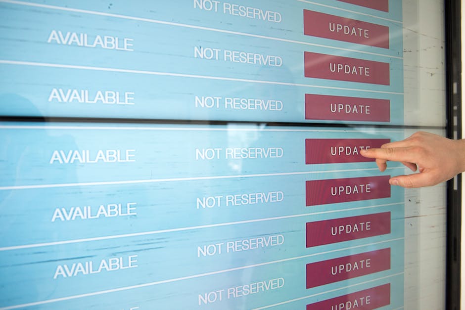 A person with a light skin tone and neatly trimmed nails is pointing at a digital display board with their index finger. The board shows multiple rows with the words 'AVAILABLE,' 'NOT RESERVED,' and r